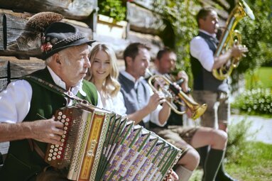 Mann in Tracht spielt steirische Harmonika, im Hintergrund eine Frau und andere Musiker. | © SalzburgerLand Tourismus
