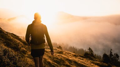 A person hikes at sunrise on a grassy hill overlooking mist-covered forests. | © Holiday Region National Park Hohe Tauern - Gerald Demolsky