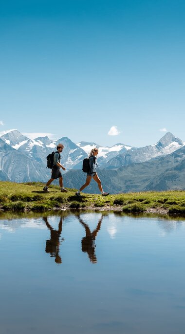 Zwei Wanderer spiegeln sich in einer Wetterlacke vor den Hohen Tauern. | © Ferienregion Nationalpark Hohe Tauern - Mathäus Gartner