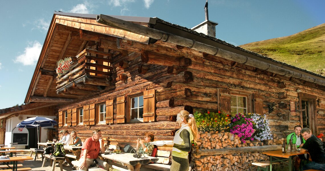 Rustikale Berghütte mit Blumen und Gästen auf der Terrasse unter blauem Himmel. | © Ferienregion Nationalpark Hohe Tauern