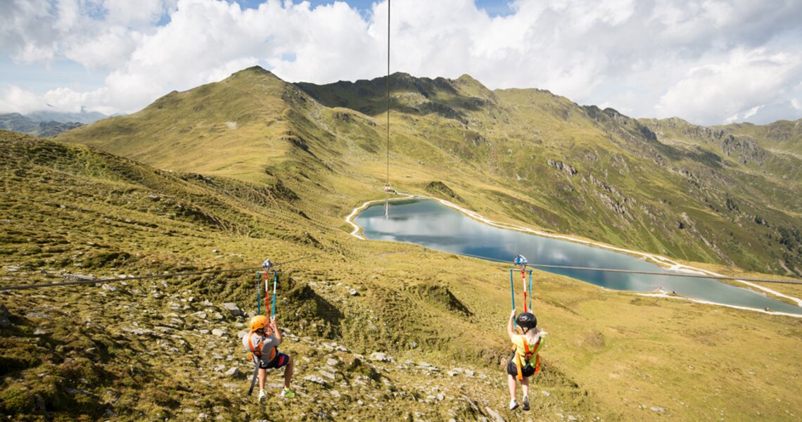 Zwei Personen auf einer Seilrutsche über einer Berglandschaft mit einem klaren Bergsee. | © Dorfbahnen Königsleiten