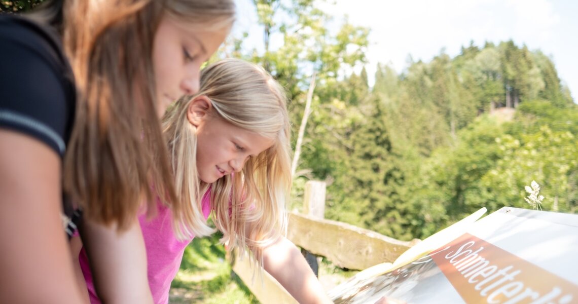 Zwei Mädchen betrachten ein Informationsschild über Schmetterlinge in einer naturnahen Umgebung. | © Ferienregion Nationalpark Hohe Tauern