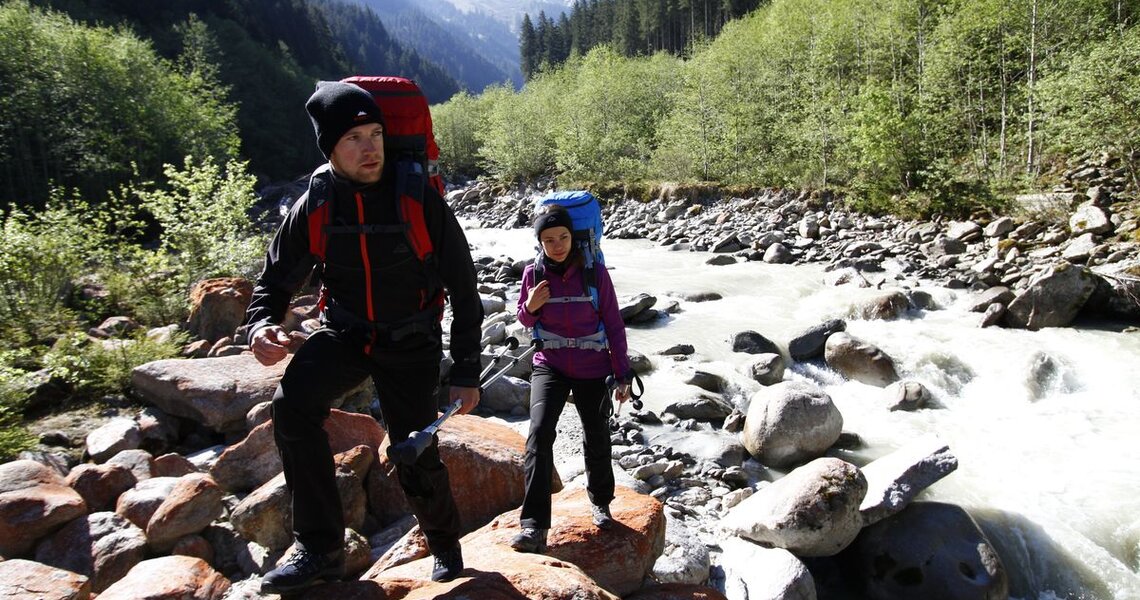 Wanderer mit Rucksäcken überqueren einen felsigen Fluss in einer alpinen Landschaft. | © Ferienregion Nationalpark Hohe Tauern