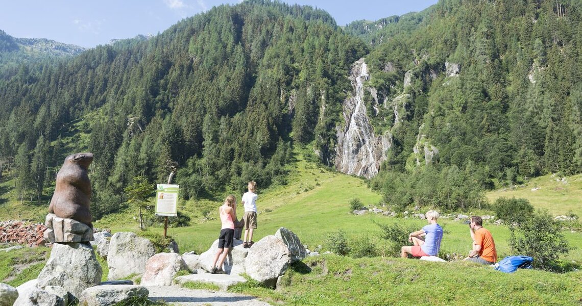 Familie beobachtet eine Murmeltierstatue und einen Wasserfall im Hintergrund. | © Ferienregion Nationalpark Hohe Tauern
