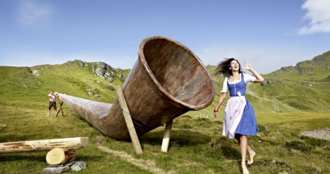 Woman in traditional dress posing next to a giant alphorn in an alpine meadow. | © TVB Wald/Königsleiten