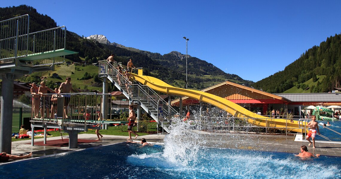 Outdoor pool with a yellow waterslide, diving platform, and visitors, surrounded by mountains and greenery. | © Freibad Großarl