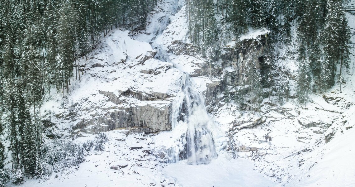 Snow-covered Krimml Waterfalls surrounded by dense fir forests with a person in the foreground. | © Daniel Breuer