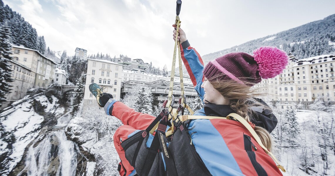 Person riding a zipline over a snowy landscape with buildings and a waterfall. | © Gasteinertal Tourismus GmbH - Klaus Listl