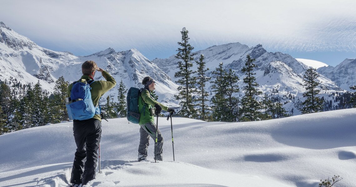 Zwei Schneeschuhwanderer genießen eine verschneite Berglandschaft mit Tannen und sonnigem Himmel. | © Helga Baldinger