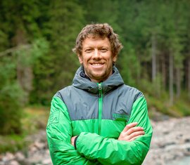 Smiling employee of the National Park Administration Hohe Tauern in Salzburg, in a National Park jacket by a stream. | © NPHT / Glück