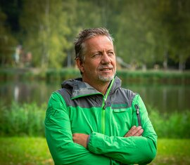 Employee of the National Park Administration Hohe Tauern in Salzburg, in a National Park jacket with crossed arms by a lake. | © NPHT / Glück