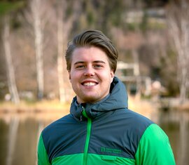 Smiling employee of the National Park Administration Hohe Tauern in Salzburg, in a National Park jacket by a lake. | © NPHT / Kreidl & Glück