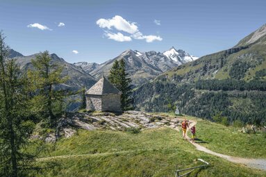 Zwei Wanderer auf einem Bergweg neben einer Kapelle vor alpiner Bergkulisse. | © Alpina Antica Trail