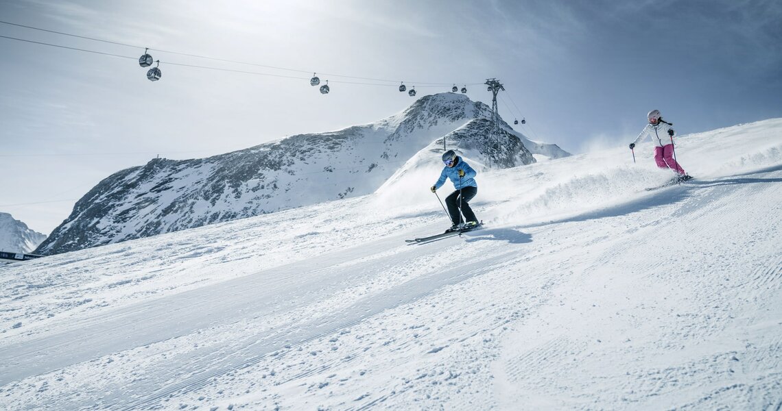 Zwei Skifahrer auf Abfahrt mit Gondeln und Berggipfeln im Hintergrund.  | © Kitzsteinhorn