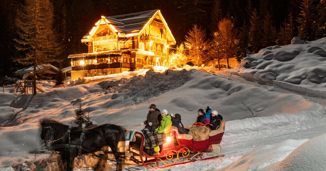 Horse-drawn sleigh ride at night in snowy winter landscape with illuminated chalet. | © Manuel Marktl