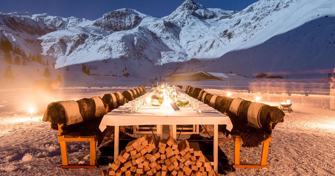 Langer, festlich gedeckter Tisch im Schnee, umgeben von Bergen und beleuchtet von Fackeln. | © Marktl Photography