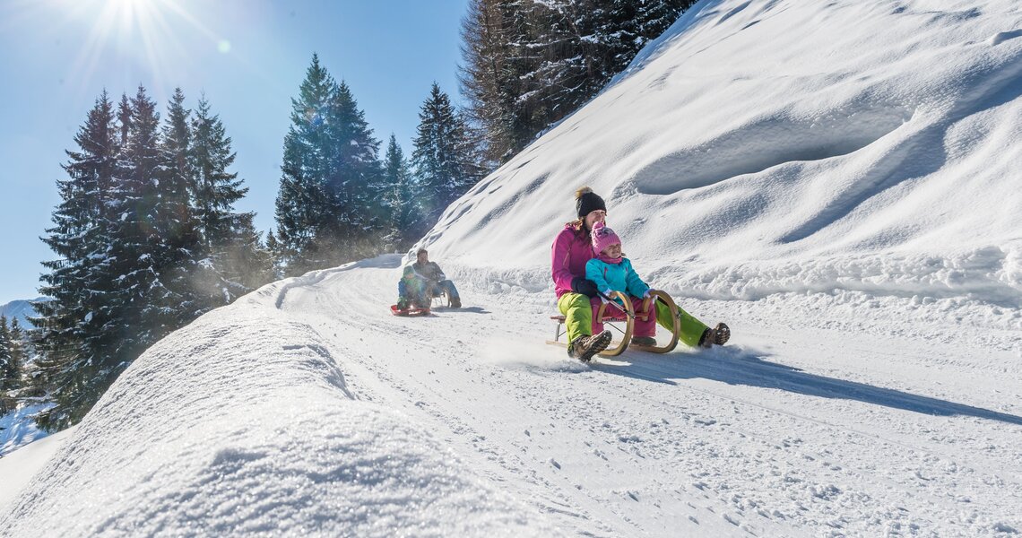 Familien rodeln bei Sonnenschein eine verschneite Winterbahn im Wald hinunter. | © Fotostudio Creatina