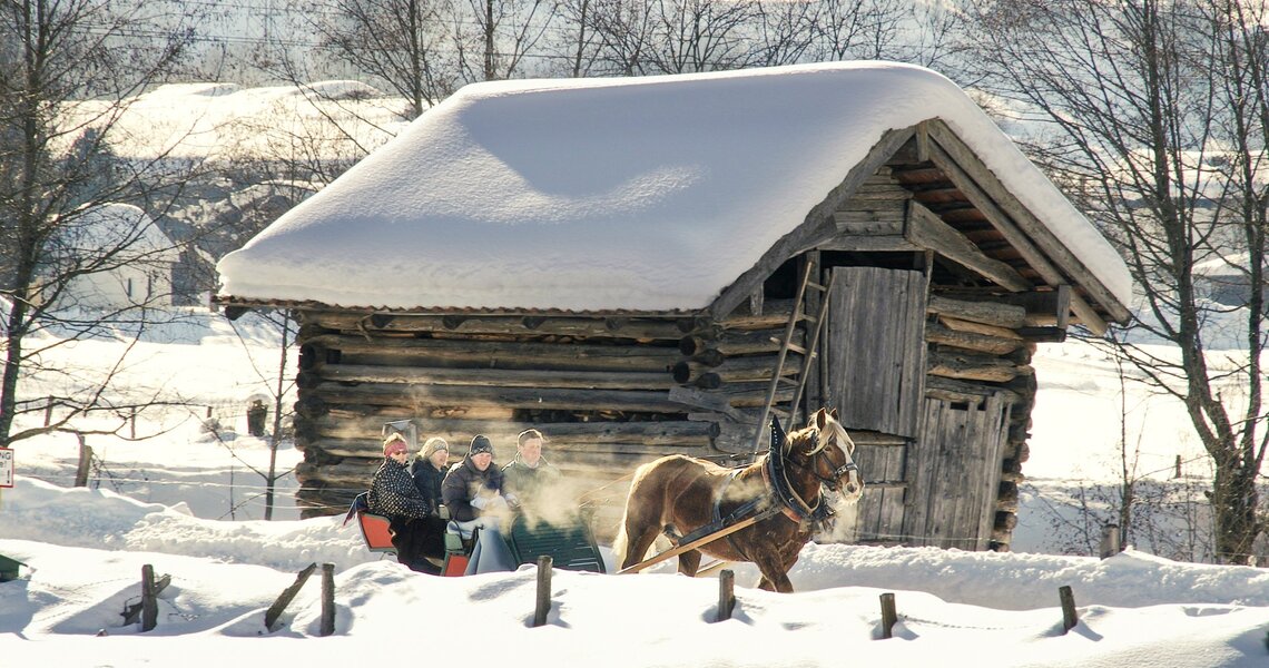 Horse sleigh with people in front of snowy wooden hut. | © Harry Liebmann