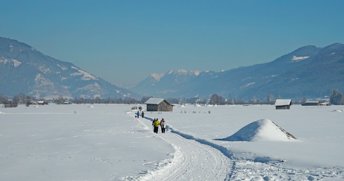 Winter hikers on a snowy path across wide fields with mountain view.