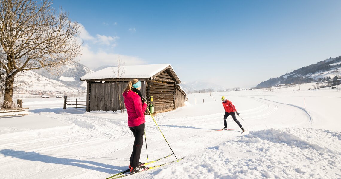 Cross-country skiers near a wooden hut in a snowy winter landscape. | © Harry Liebmann
