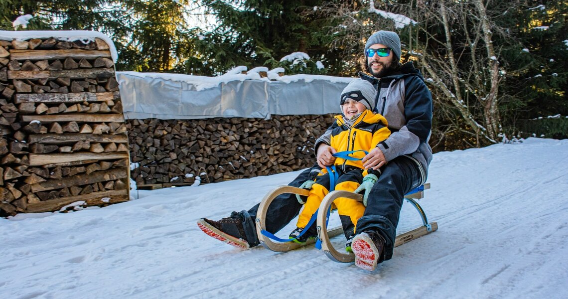 Father and child laughing while sledding down a snowy path.
