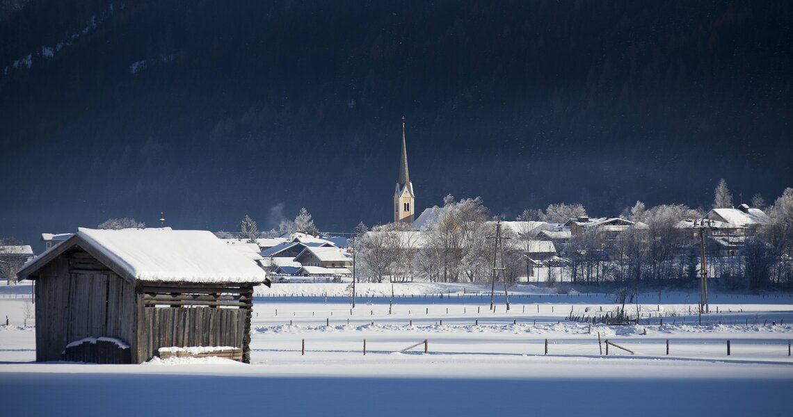 Verschneite Landschaft mit Holzhütte und Kirchturm im Hintergrund. | © Franz Reifmüller