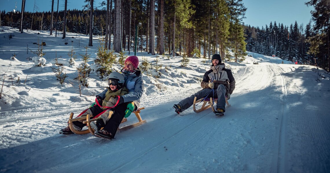 Family happily sledding side by side down a snowy forest trail. | © Sobietzki