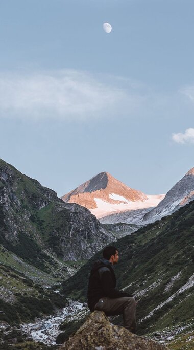 Alpine valley at sunrise with the moon in the sky and a hiker in the foreground. | © Hohe Tauern Nationalpark - Tobias Kaser